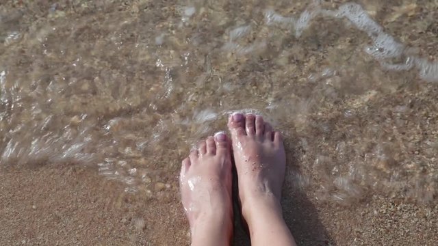 Closeup top view video of female feet with fresh french pink and white pedicure. Woman relaxing at summer sandy beach sitting on seashore near sea water. Real time point of view  footage.