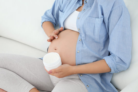 Pregnant woman applying body cream on belly at home, closeup