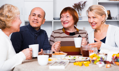 Smiling aged friends are sitting at table with tea and talking about life
