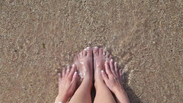 Closeup top view video of female feet and hands with fresh french pink and white pedicure and manicure. Woman relaxing at summer sandy beach sitting on seashore near sea water. Real time footage.