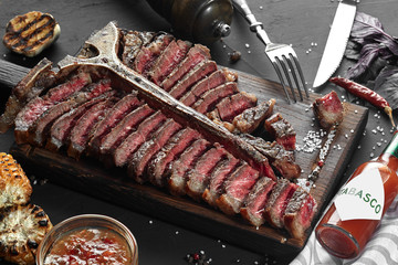 Sliced Steak T-bone lying on wooden board. Grilled vegetables in a pan grill. Black table with gray cloth