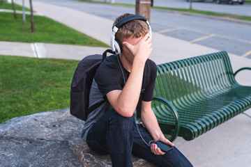 Ashamed teenager sitting on a rock outside of a school while listening to music.