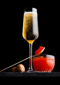 Elegant glass of yellow champagne with red caviar on golden spoon and glass container of caviar on marble board on black background.