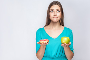 Portrait of dreaming young beautiful girl in blue blouse standing, holding pink donut and green apple in the hands like weighing with thoughful face on grey background. Indoor, isolated,copy space