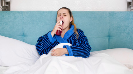 Portrait of sick woman with sore throat lying in bed and using throat spray