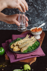 Chef's hands sprinkle with seasoning Fried fish in a plate with  tomato sauce, greens and peppers on wooden board  and a dark background