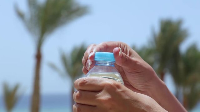 Closeup view of tanned female hands opening closed plastic bottle with fresh clear water isolated at blurry tropical beach background with blue sky and green palms. Real time full hd video footage.
