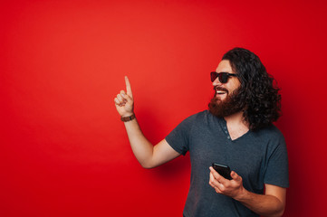 Smiling happy bearded man holding smartphone and pointing away at copyspace over red background