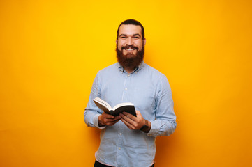 Cheerful bearded man in casual holding a book and looking confident at the camera on yellow background