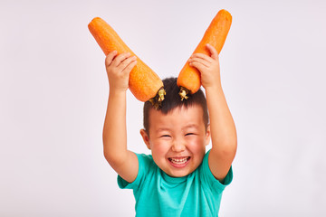 Child boy in turquoise shirt, holds huge carrots depicting horns - fruits and healthy food