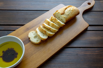 A french bread baguette sliced up on a cutting board with a bowl of olive oil with a splash of balsamic vinegar along side a bottle of olive oil that had a stem of rosemary.