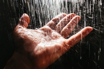 A man's hand in a spray of water in the sunlight against a dark background. Water as a symbol of purity and life