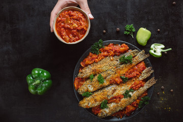Fried fish in a plate with  tomato sauce, greens and peppers on dark background