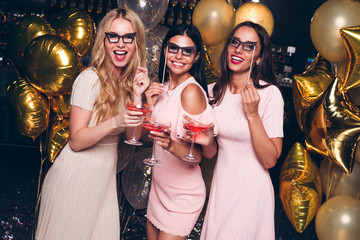Pink party. Beautiful young women in pink dresses holding eyeglasses and looking at camera with smile while celebrating in nightclub