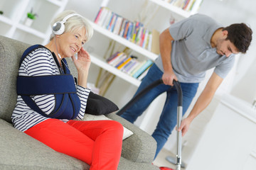 senior female sitting on sofa while home help is cleaning