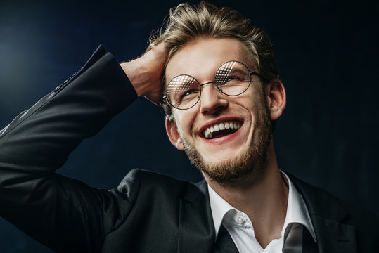Close up studio portrait of young confident handsome happy smiling man wearing round glasses, white shirt, black jacket, touching his hair, looking up, posing on dark background