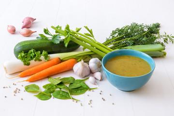 Turquoise bowl with spinach soup and raw ingredients, spice and herbs. The spinach leafs, onion shallots, leeks, celery, zucchini, garlic, parsley, carrot and white pepper, white wooden background.