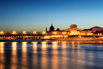 Obraz na płótnie Canvas Illuminated Chain bridge reflecting in Danube river, silhouette of Parliament domes