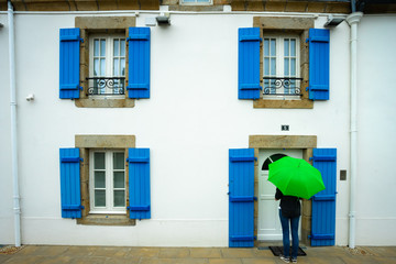 une personne avec un parapluie vert devant une façade de maison blanche avec des fenêtres aux volets bleus