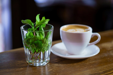 transparent glass with a fresh mint inside stands on the background of a cup with a latte in the cafe. Close-up