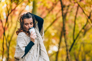 a pensive woman stands with a cup of coffee in the park and straightens her curly hair on the background of autumn park. Close-up