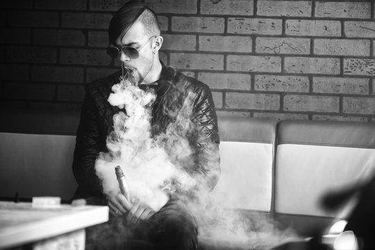 Vaping teenager. Young handsome bearded white man in a black jacket and sunglasses is smoking an electronic cigarette and letting off puffs of steam in vape bar. Bad habit. Black and white.