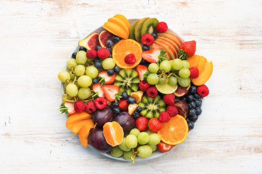 Healthy fruit platter, strawberries raspberries oranges plums apples kiwis grapes blueberries on the white wooden table, top view, copy space for text, selective focus