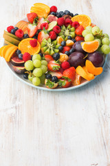 Healthy fruit platter, strawberries raspberries oranges plums apples kiwis grapes blueberries on the white wooden table, vertical, copy space for text, selective focus