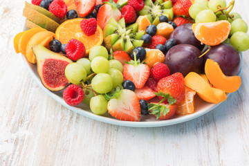 Healthy fruit platter, strawberries raspberries oranges plums apples kiwis grapes blueberries on the white wooden table, copy space for text, selective focus