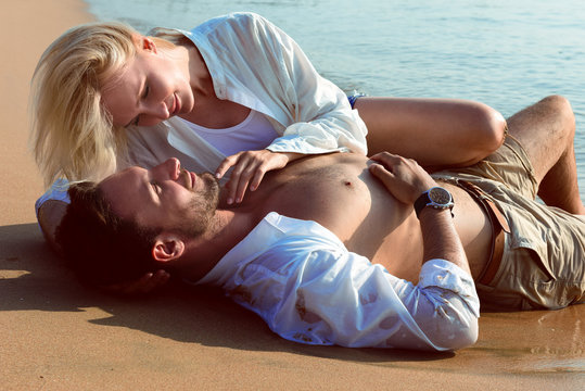 Beautiful happy couple in love lying on a beach on sand in a white shirts are in love smiling. Handsome bearded men and sexy blonde women in water in wet clothes having fun kissing, and laughing