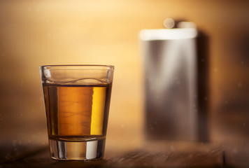 Whiskey shot glass on top of a wooden table background and a metal hip flask in the background