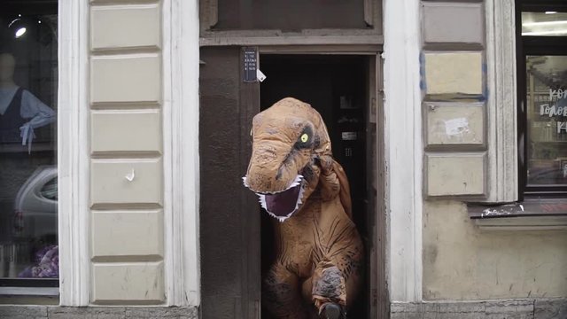 Person in t rex mascot costumeout city house metal door entrance at street on sunny day, slow motion
