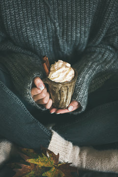 Woman in dark woolen sweater, grey jeans and white woolen socks sitting and holding mug with hot chocolate or coffee with whipped cream and cinnamon sticks in hands. Fall warming sweet drink
