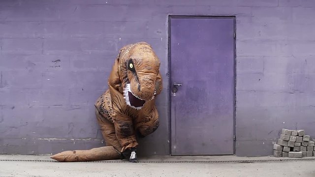 Person in t rex mascot costume cross arms standing leans next to door on purple wall at street on sunny day, slow motion