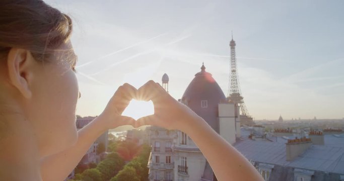 young woman hands making heart shape gesture holding sun flare enjoying romantic travel vacation in paris france looking at beautiful eiffel tower