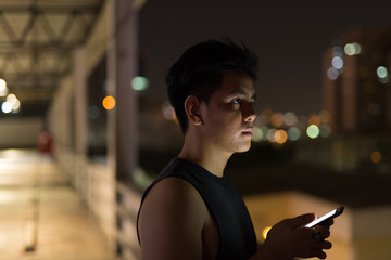 Young handsome Asian man thinking while using phone against view of the city at night