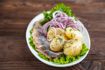 Russian traditional cuisine on a dark textured wooden background. Potatoes with dill and herring with onions and greens on a white plate.