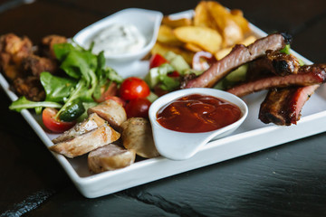 Meat plate with pork ribs, chicken, fried potatoes, cheese balls, croutons, sausages with sauce on black background
