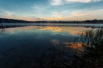 Beautiful, red dawn on the lake. The rays of the sun through the fog. The blue sky over the lake, the morning comes, the sky is reflected in the water.