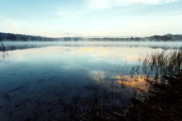 Beautiful, red dawn on the lake. The rays of the sun through the fog. The blue sky over the lake, the morning comes, the sky is reflected in the water.