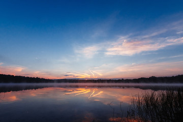 Beautiful, pink violet dawn over the lake. Fog over the lake, the rays of the sun, very dense fog, dawn, the blue sky over the lake, the morning comes, the forest reflects in the water.