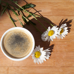 A cup of black coffee with foam and  bouquet of white camomile flowers on  wooden background in sun light with  shadow top view 