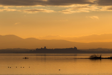 Beautiful view of Trasimeno lake (Umbria, Italy) at sunset, with orange tones, birds on water, a man on a canoe and Castiglione del Lago town on the background