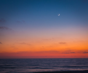 As the sun goes down and seems to melt into the ocean, the skies darken a bit and the moon begins to shine in the seaside sky as friends and lovers sit and enjoy the scene and each other