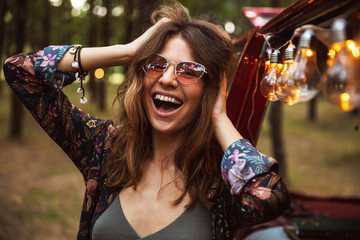 Image of young hippy woman, wearing stylish accessories smiling while resting in forest camp