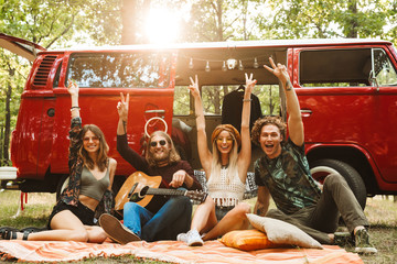 Group of friends hippies men and women laughing, and sitting near vintage minivan into the nature