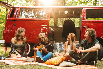 Group of friends hippies men and women playing guitar, and sitting near vintage minivan into the nature