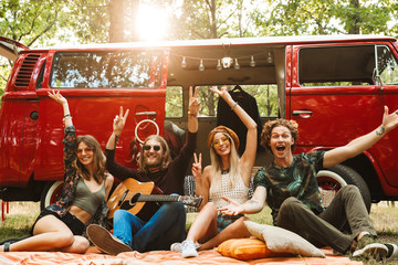 Group of young hippies men and women laughing, and sitting near vintage minivan into the nature