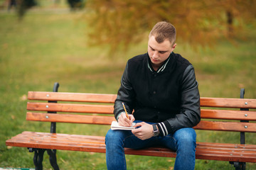 Student in a black jacket sits in a park on a bench writes down his thoughts in a notebook. Handsome boy