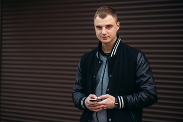A young guy in a black jacket against a background of dark striped walls uses the phone